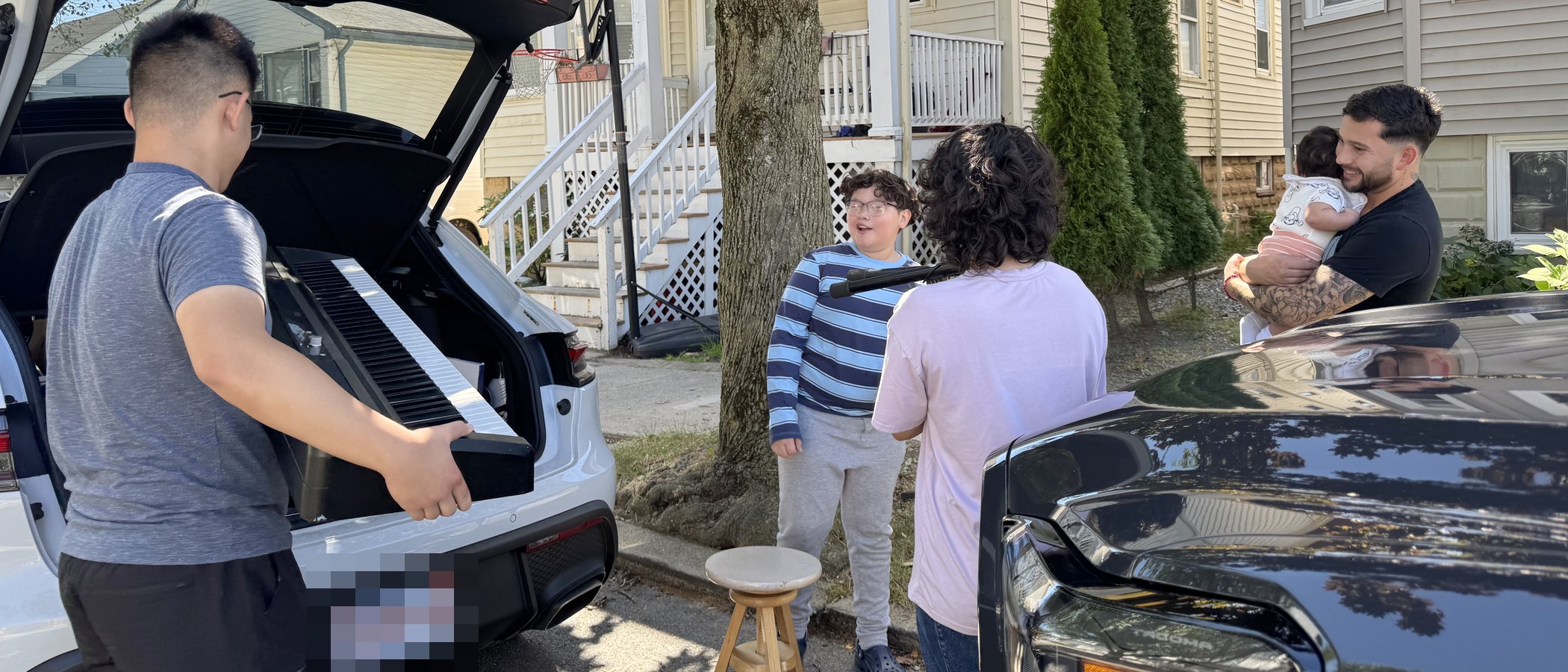 A student lifting a donated digital piano off the tailgate, outside the house it is going to.