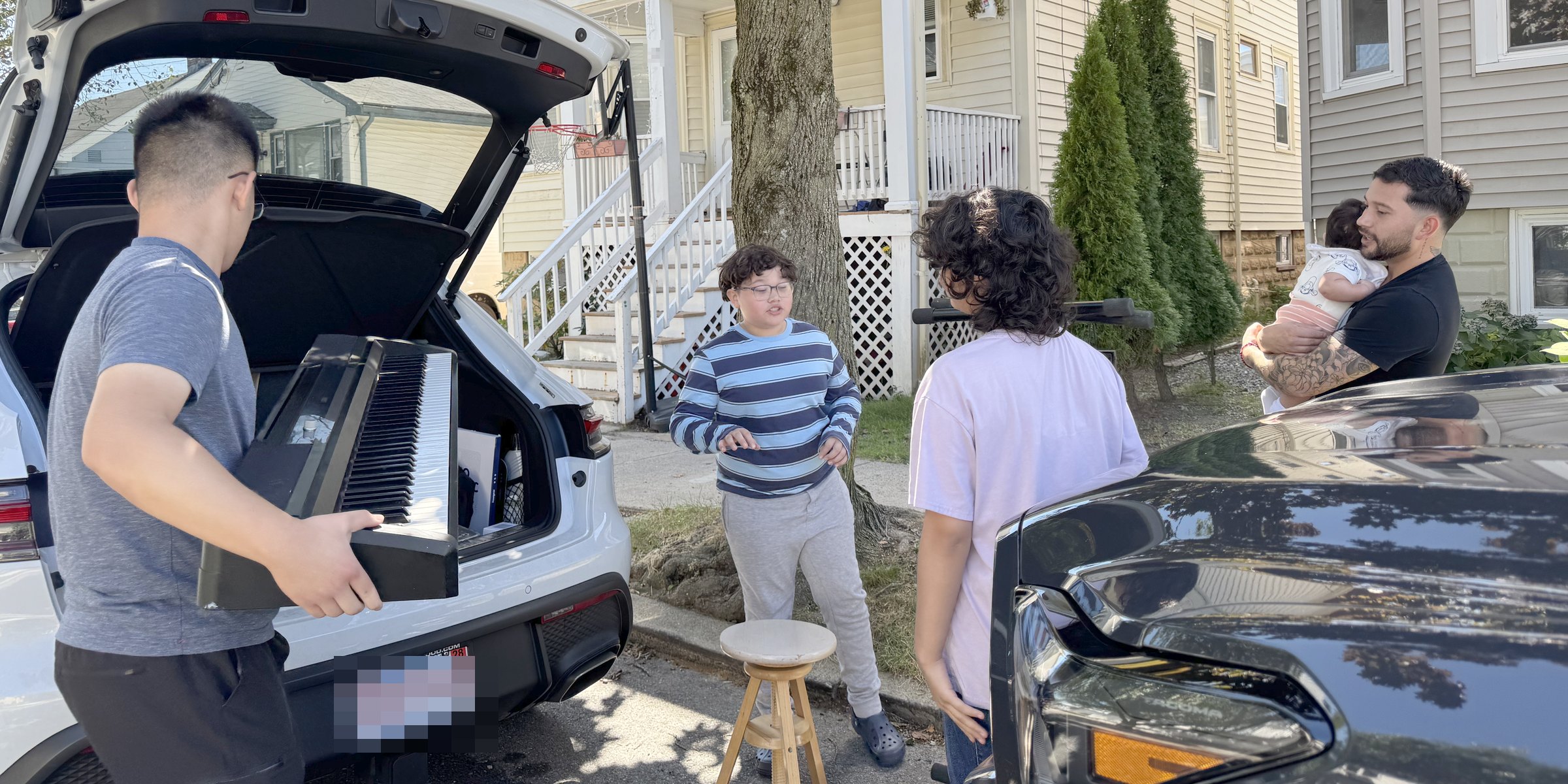 A student lifting a digital piano out of a car while the family who asked for it waits on the sidewalk.