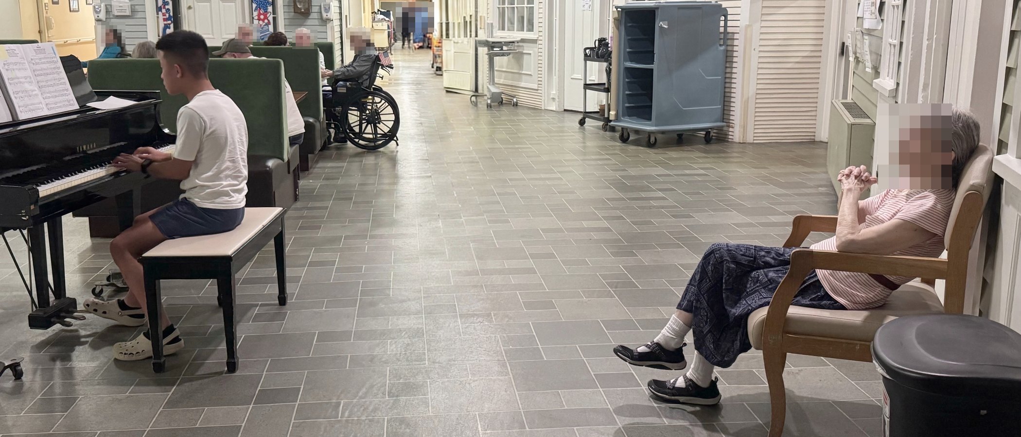 A student playing a grand piano at one end of a residence hallway, with a woman listening from a chair at the other.